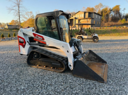 White Bobcat skid-steer loader with a large bucket resting on a gravel lot; residential construction site in the background, including a two-story house and a golf cart nearby.