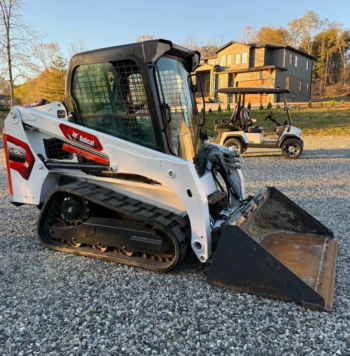 White Bobcat skid-steer loader with a large bucket resting on a gravel lot; residential construction site in the background, including a two-story house and a golf cart nearby.