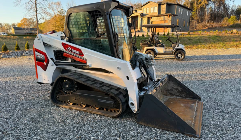 White Bobcat skid-steer loader with a large bucket resting on a gravel lot; residential construction site in the background, including a two-story house and a golf cart nearby.